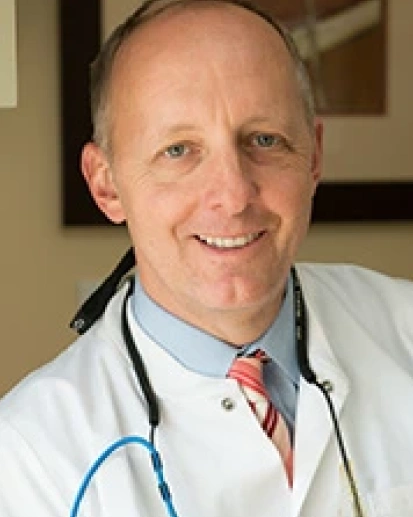 Male healthcare professional in white coat with stethoscope smiling at camera in medical office.