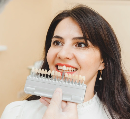 Woman holding dental shade guide against teeth, smiling in dental office setting.