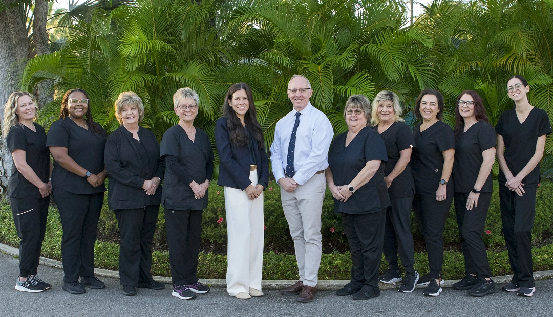 Professional dental team of twelve staff members in black uniforms standing outdoors with green foliage background.