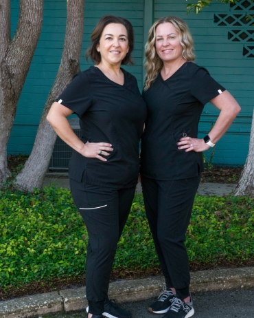 Two women in black athletic wear pose together outdoors against turquoise wall and greenery.