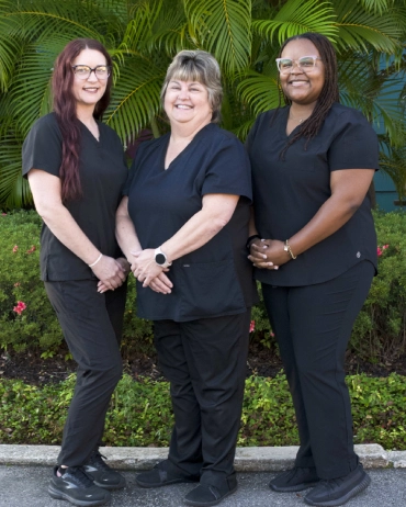 Three dental professionals in black scrubs posing together indoors with green plants in background.