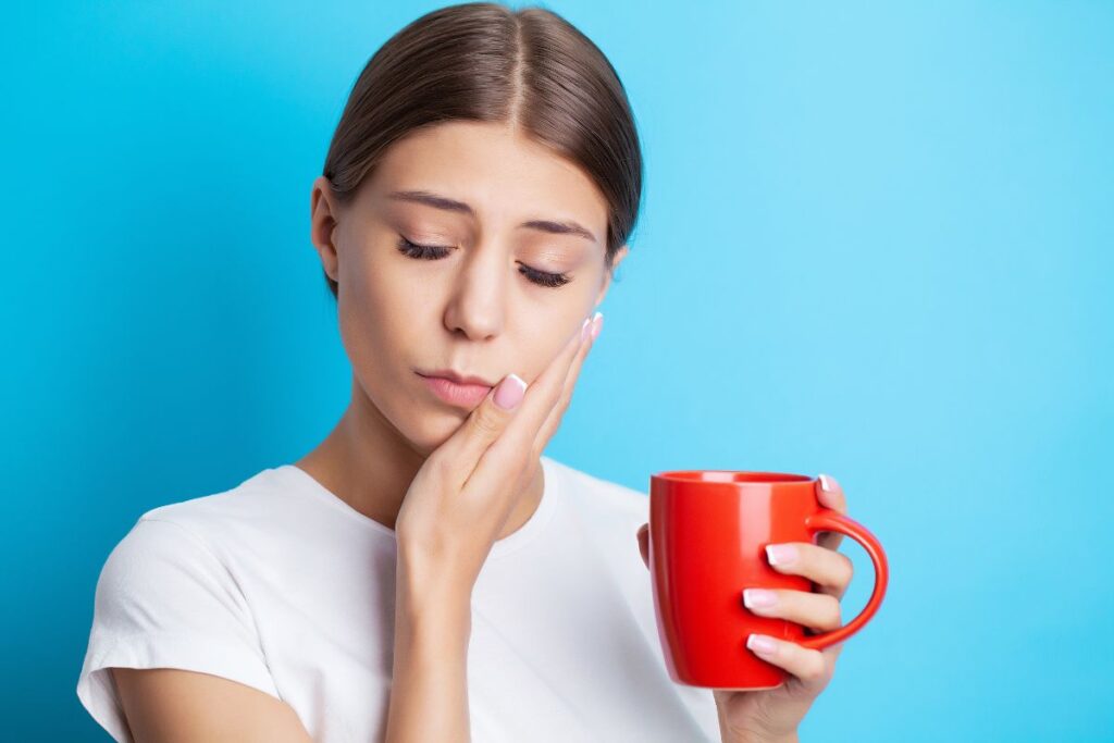 A woman holds a cup while touching her cheek in discomfort, illustrating tooth sensitivity that can be triggered by sugary or acidic beverages.