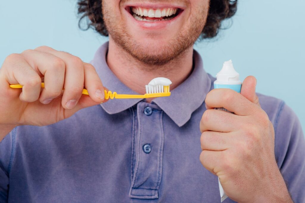 Smiling man holding a yellow toothbrush with toothpaste applied while demonstrating proper oral hygiene and daily dental care routine against a light blue background.